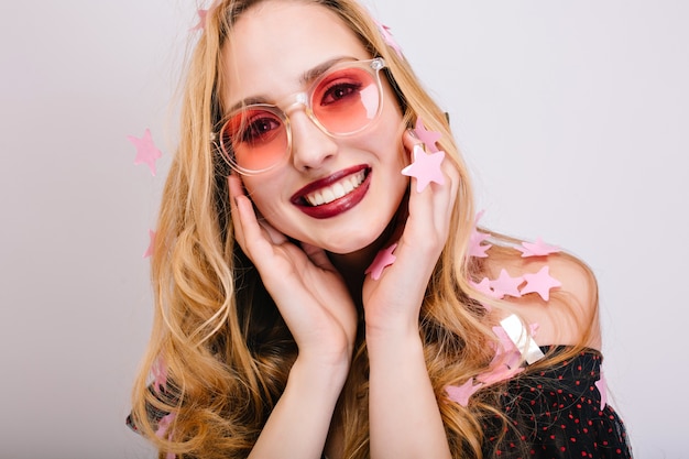 Closeup retrato de jovem loira sorridente de óculos cor de rosa, sessão de fotos de festa, confetes em todos os lugares. Tem um sorriso lindo, cabelos longos e encaracolados, fica legal no vestido preto.