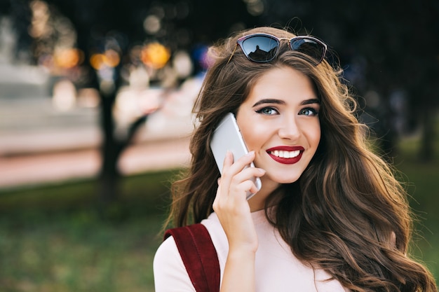 Closeup retrato de linda garota com maquiagem eficaz e cabelos longos, falando no telefone no parque. Ela tem lábios carnudos e sorridentes.