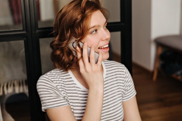 Closeup retrato de menina encaracolada vermelha em t-shirt listrada, falando no telefone.