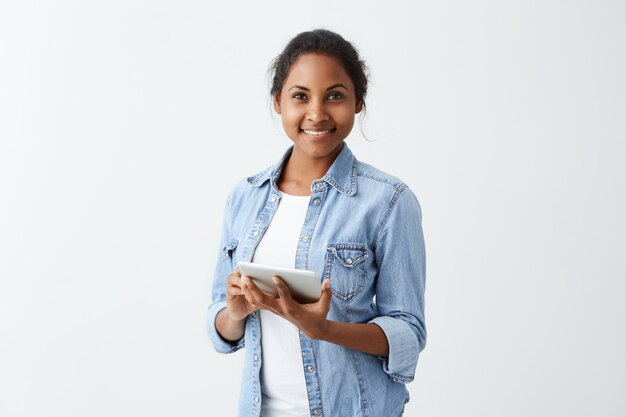 Closeup retrato de mulher de pele escura, segurando o tablet nas mãos, sorrindo com sorriso perfeito e dentes brancos na parede branca isolada. Emoção humana positiva facial ex