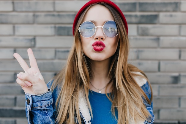 Closeup retrato de mulher incrível com expressão de rosto beijando em frente a parede de tijolo foto ao ar livre de alegre jovem de óculos e jaqueta jeans posando na rua