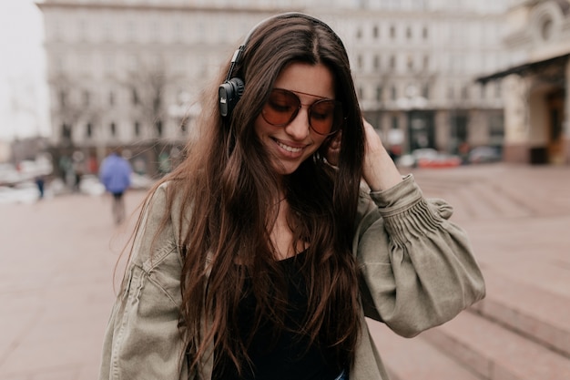 Closeup retrato de mulher sorridente alegre relaxando com música na cidade. Foto ao ar livre de modelo feminino sorridente em fones de ouvido, passando o fim de semana