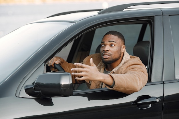 Closeup retrato de nervoso jovem negro sentado em seu carro. Homem com barba, vestindo jaqueta marrom. Motorista masculino parecendo nervoso.