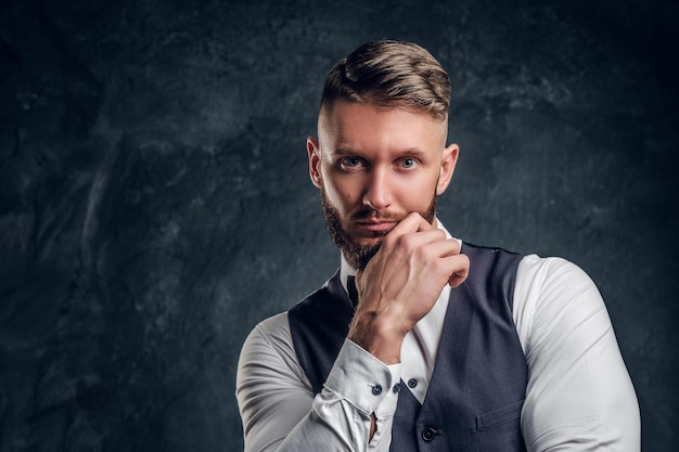 Closeup retrato de um jovem elegantemente vestido com elegante barba e cabelo segurando a mão no queixo e olhando para uma câmera. foto de estúdio contra o fundo da parede escura