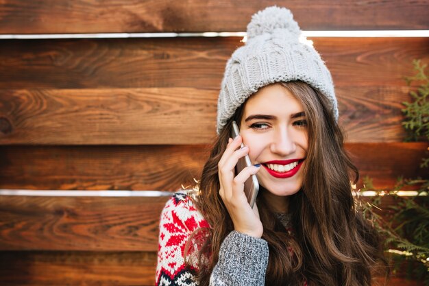 Closeup retrato linda garota com cabelo comprido e sorriso branco de neve com chapéu de malha na madeira. Ela usa um suéter quente, falando ao telefone, sorrindo para o lado.