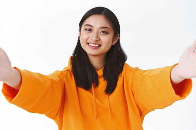 Closeup retrato tenra jovem asiática com capuz laranja elegante bem-vindo alguém segurando a câmera esticando as mãos para o lado para abraçar ou abraçar a pessoa mais querida fundo branco
