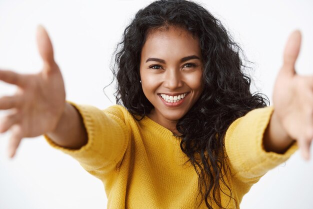 Closeup tenra e bonita mulher de cabelos encaracolados afro-americana em suéter amarelo esticando as mãos para a frente sorrindo e olhando para a câmera com amor e cuidado para dar abraço abraço amigo