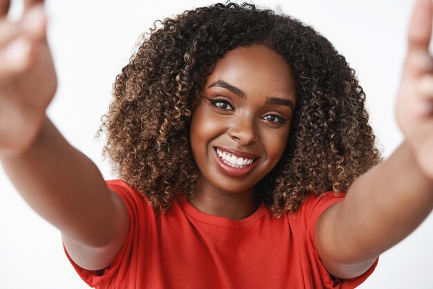 Closeup tiro de linda mulher afro-americana delicada e feminina com cabelos cacheados em camiseta vermelha, puxando as mãos e segurando a câmera como abraço ou amigo reconfortante com um sorriso suave no rosto