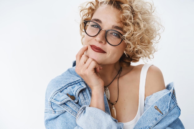 Closeup tiro de menina moderna romântica e autoconfiante com penteado loiro e óculos sorrindo sedutor e terno segurando os dedos na mandíbula olhando bonito para a câmera sobre fundo branco