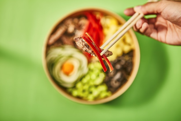 Closeup tiro de uma mão humana segurando um pedaço de carne com pimenta vermelha perto de palitos chineses