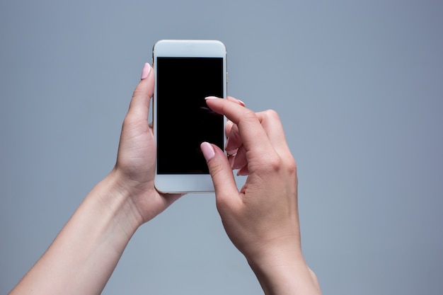 Closeup tiro de uma mulher digitando no celular em fundo cinza. Mãos femininas segurando um smartphone moderno e apontando com o dedo.