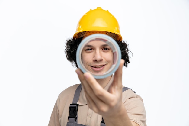 Closeup vista sorridente jovem trabalhador da construção civil vestindo uniforme e capacete de segurança esticando a fita adesiva para a câmera olhando para a câmera através dela isolada no fundo branco