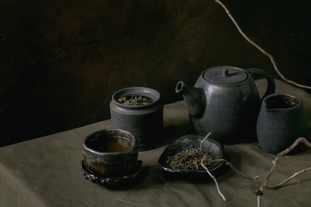 Coleção de chaleira de bule de cerâmica preta artesanal artesanal, jarro, jarra, xícara wabi sabi, pires para a cerimônia do chá em pé na toalha de mesa de linho com galho seco em quarto escuro.