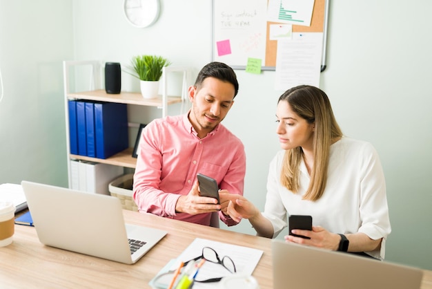 Colega de trabalho latino masculino mostrando um e-mail de trabalho em seu smartphone para sua colega caucasiana enquanto está sentado em sua mesa de escritório