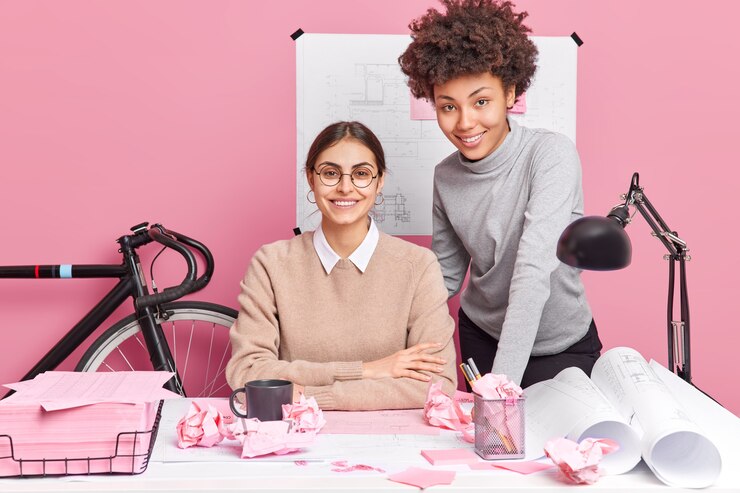 Foto grátis colegas de mulheres felizes, preparadas para a sessão de trabalho, têm expressões alegres pose na área de trabalho estando de bom humor pose na mesa do escritório rodeado por desenhos de projetos de papéis. conceito de trabalho em equipe