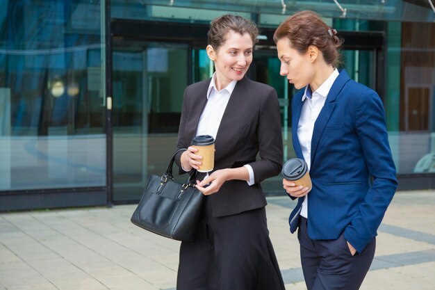 Colegas de trabalho do sexo feminino alegres bebendo café ao ar livre, sorrindo, rindo. Jovens empresárias de terno, caminhando juntas na cidade. Conceito de pausa para o trabalho