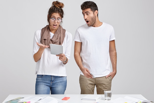 Foto grátis colegas de trabalho femininas e masculinas terríveis e surpresas olham com expressão estupefata para o bloco de notas, recebem comentários e observações ruins sobre seu trabalho de projeto, ficam perto da parede branca perto da mesa