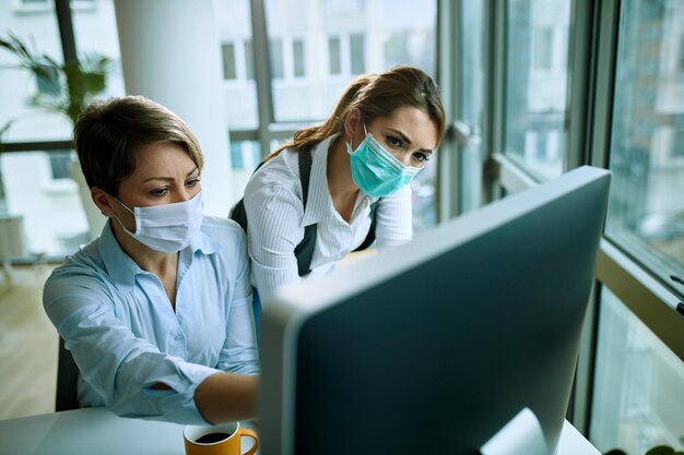 Colegas de trabalho femininas usando máscaras faciais e cooperando enquanto usam PC de mesa no escritório