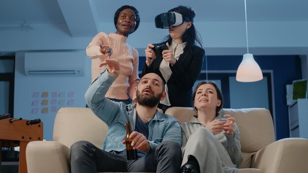 Colegas de trabalho usando a tecnologia para jogar videogame depois do trabalho. Colegas jogando com óculos de realidade virtual e controle na televisão, desfrutando de bebidas e lanches depois do expediente para se divertir.