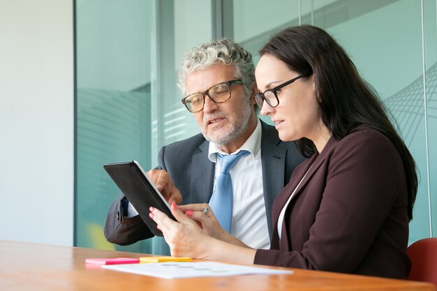 Colegas masculinos e femininos usando tablet juntos, olhando e apontando para a tela do gadget enquanto está sentado à mesa no escritório.