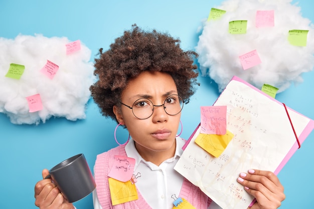 Foto grátis colegial seriamente zangada e descontente com cabelo afro se prepara para a prova de matemática faz anotações tenta se lembrar de fórmulas bebidas café usa óculos redondos poses contra adesivos azuis de parede ao redor