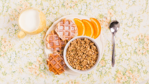 Colher; jarro de leite e prato de tigela de granola saudável com waffles e fatia de uma laranjas sobre fundo floral