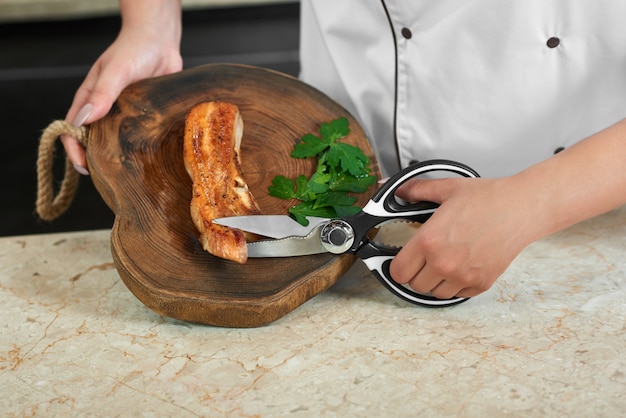 Colhido perto acima de um cozinheiro chefe fêmea que corta a carne grelhada com as tesouras que trabalham na cozinha do restaurante.