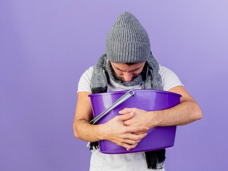 Foto grátis com a cabeça baixa, jovem doente usando um chapéu de inverno com lenço segurando uma cesta de plástico e vomitando dentro dela, isolado no fundo roxo