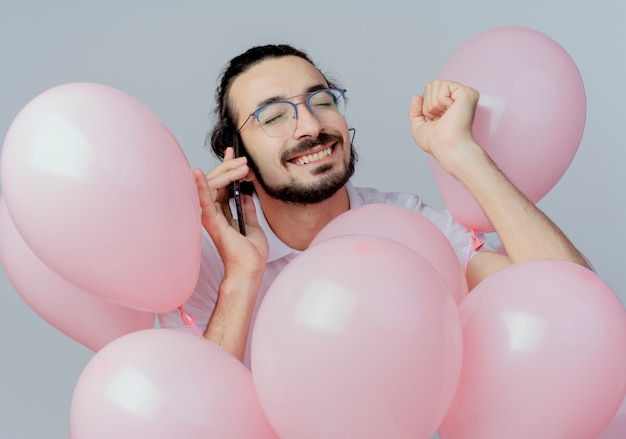 Com olhos fechados alegre homem bonito de óculos em pé entre balões e fala ao telefone isolado no branco