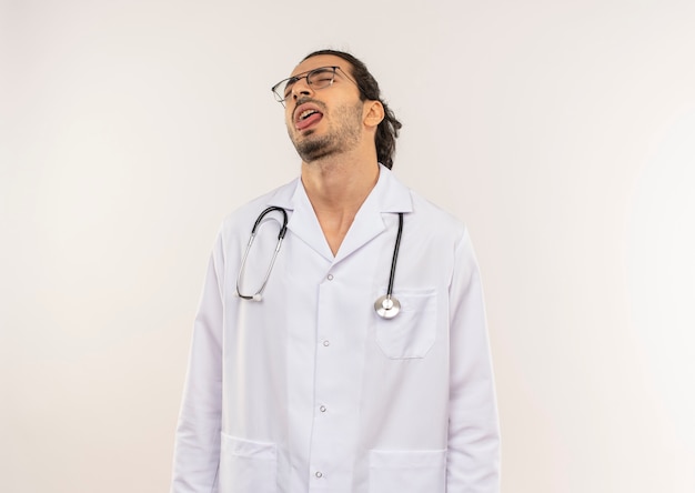 Com olhos fechados, jovem médico do sexo masculino com óculos ópticos, vestindo túnica branca com estetoscópio mostrando a língua na parede branca isolada com espaço de cópia