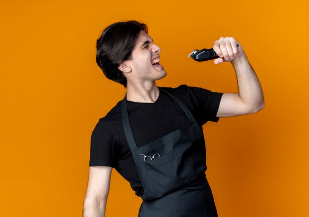 Com os olhos fechados, alegre jovem barbeiro bonito de uniforme segurando uma tesoura de cabelo e cantando isolado em laranja