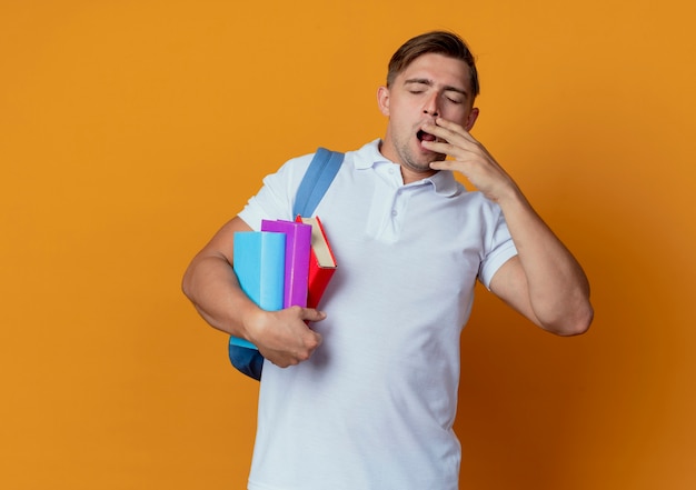 Com os olhos fechados, bocejando jovem e bonito estudante do sexo masculino usando uma bolsa nas costas segurando livros