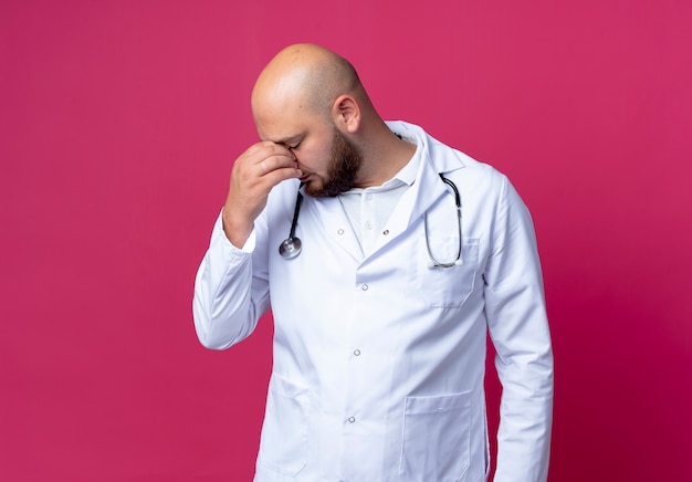 Com os olhos fechados, cansado jovem médico careca vestindo roupão médico e estetoscópio colocando a mão no nariz isolado em rosa com espaço de cópia