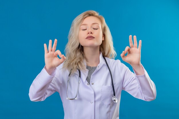 Com os olhos fechados, doutor, jovem, usando estetoscópio, em bata médica, mostrando o gesto certo sobre fundo azul