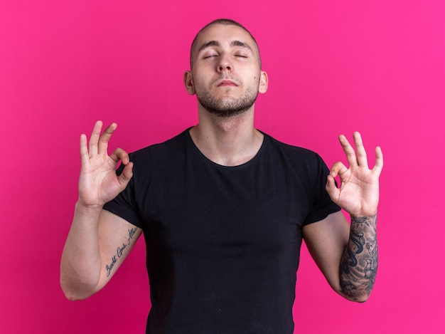 Com os olhos fechados, jovem bonito vestindo uma camiseta preta e mostrando a meditação isolada no fundo rosa