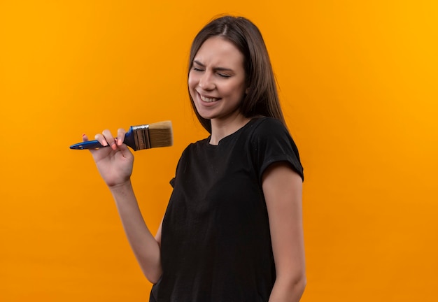 Com os olhos fechados, jovem caucasiana vestindo uma camiseta preta segurando um pincel e fingindo que canta na parede laranja isolada