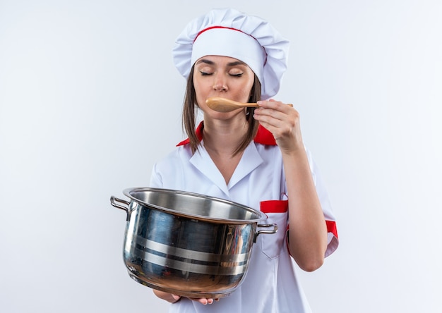 Com os olhos fechados, jovem cozinheira vestindo uniforme de chef segurando uma panela, tentando sopa com uma colher isolada no fundo branco