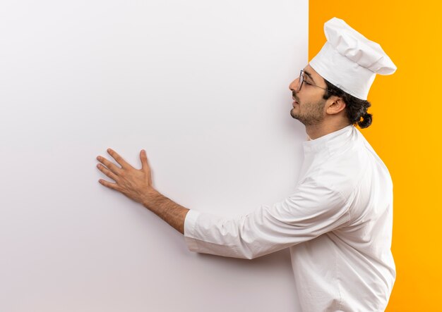 Foto grátis com os olhos fechados, jovem cozinheiro usando uniforme de chef e óculos segurando uma parede branca isolada em uma parede amarela com espaço de cópia