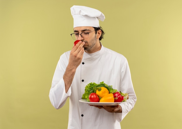 Com os olhos fechados, jovem cozinheiro vestindo uniforme de chef e óculos, segurando vegetais no prato e cheirando tomate na mão, isolado na parede verde com espaço de cópia
