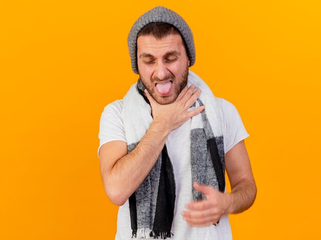 Com os olhos fechados, jovem doente usando chapéu de inverno e lenço agarrou a garganta dolorida isolada no amarelo