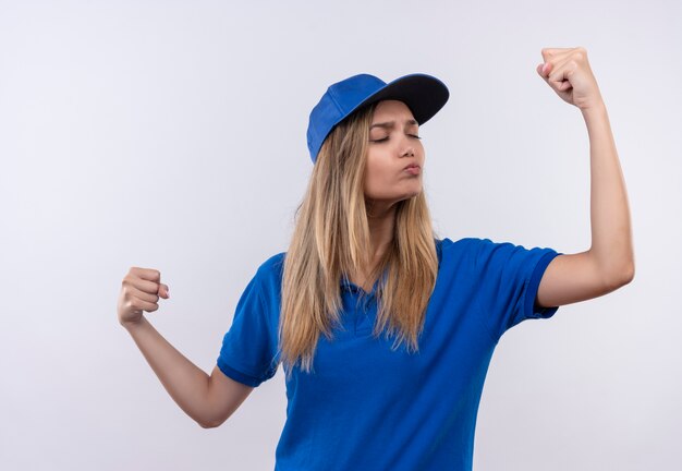 Com os olhos fechados, jovem entregadora vestindo uniforme azul e boné mostrando forte gesto isolado na parede branca com espaço de cópia