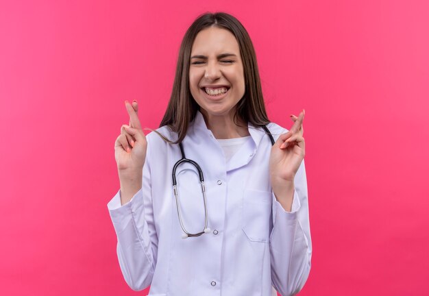 Com os olhos fechados, jovem médica usando estetoscópio jaleco cruzando os dedos na parede rosa isolada