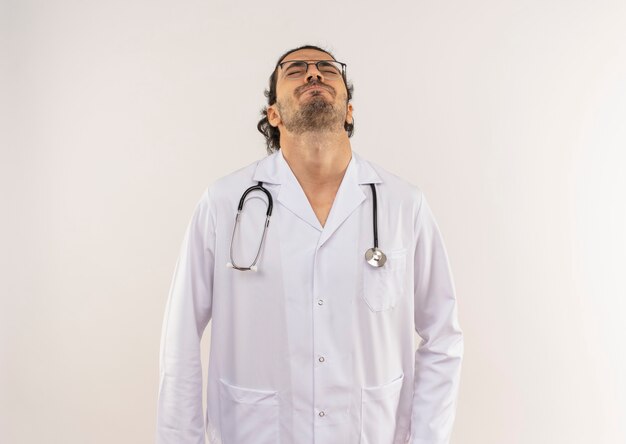 Com os olhos fechados, jovem médico do sexo masculino com óculos ópticos vestindo túnica branca com estetoscópio levantando a cabeça na parede branca isolada com espaço de cópia