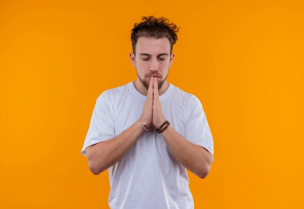 Com os olhos fechados, jovem vestindo uma camiseta branca mostrando um gesto de oração em uma parede laranja isolada