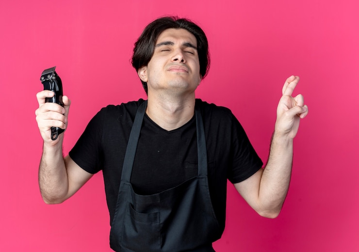 Foto grátis com os olhos fechados preocupado, jovem barbeiro bonito de uniforme segurando uma tesoura de cabelo e cruzando os dedos isolados na rosa