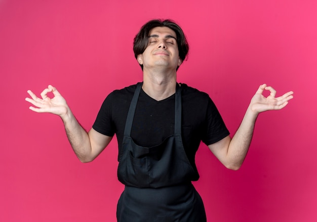 Com os olhos fechados satisfeito, jovem barbeiro bonito de uniforme fazendo meditação isolada em rosa