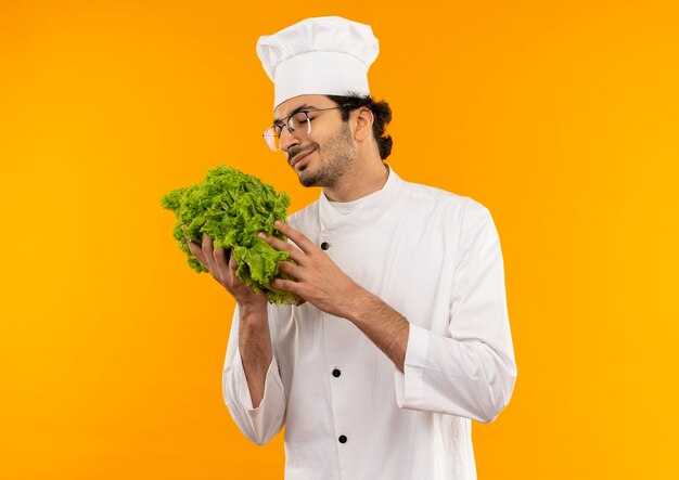 Com os olhos fechados satisfeito, jovem cozinheiro vestindo uniforme de chef e óculos segurando uma salada isolada na parede amarela