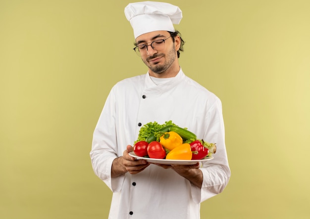 Com os olhos fechados satisfeito, jovem cozinheiro vestindo uniforme de chef e óculos segurando vegetais no prato isolado na parede verde