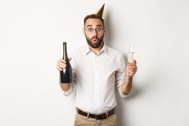 Comemoração e feriados. Homem surpreso com um chapéu de aniversário, segurando uma taça e champanhe e parecendo surpreso, em pé