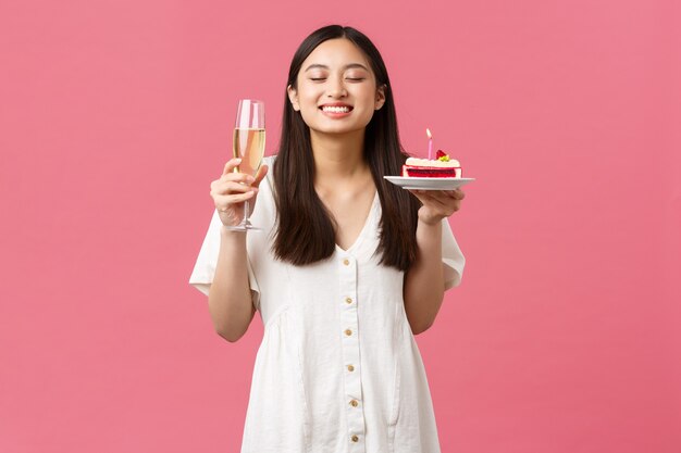 Comemoração, festas de fim de ano e conceito divertido. Uma linda mulher sonhadora comemorando seu aniversário com taça de champanhe e bolo de aniversário, sorrindo e fazendo desejo sobre fundo rosa.